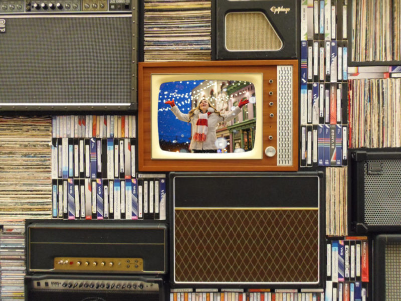 En la foto se ve una televisión antigua con una foto de navidad, rodeada de cintas de vídeo, radios, vinilos y otras antiguedades